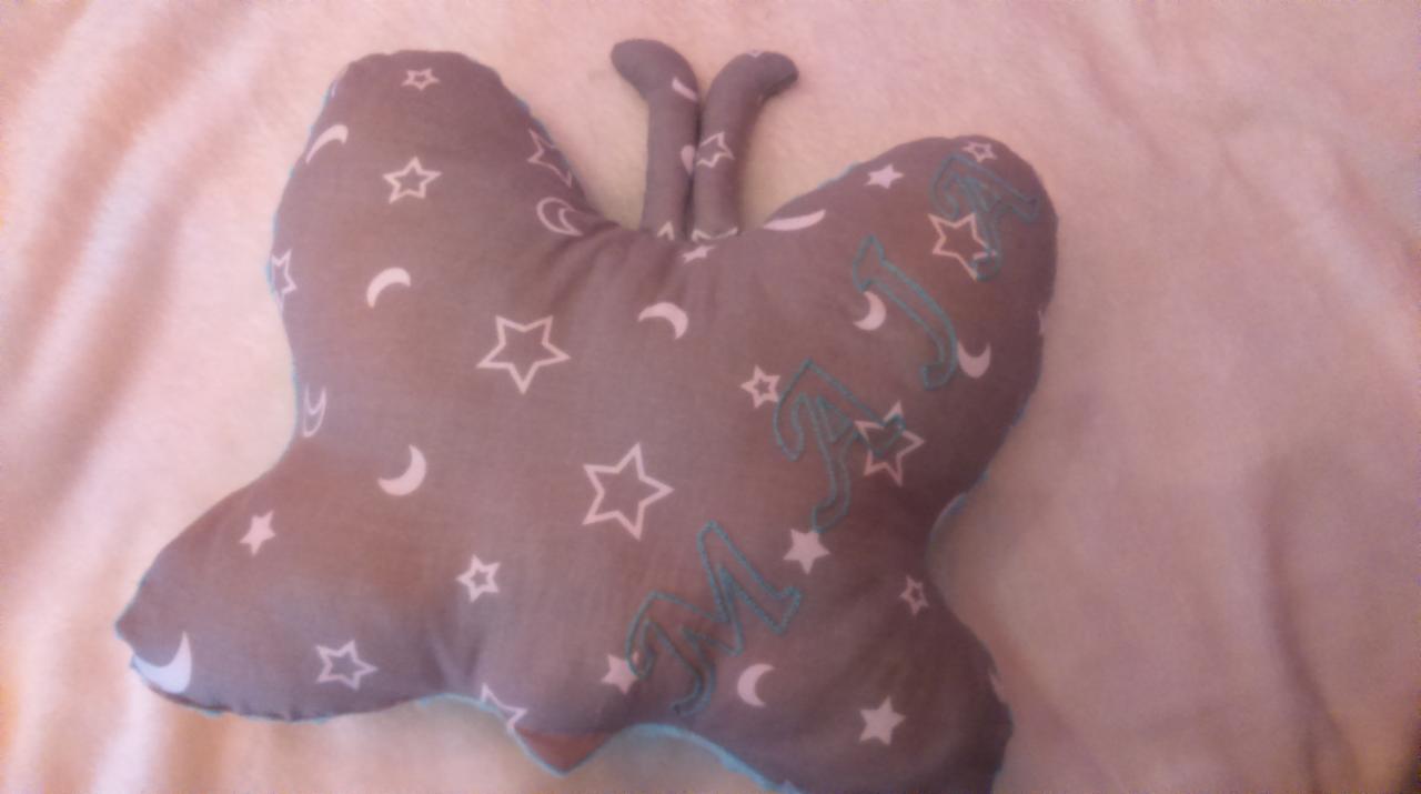 Szara poduszka w kształcie motyla, z białymi gwiazdkami i księżycami oraz wyhaftowanymi turkusowymi inicjałami, leżąca na białym, puszystym tle.