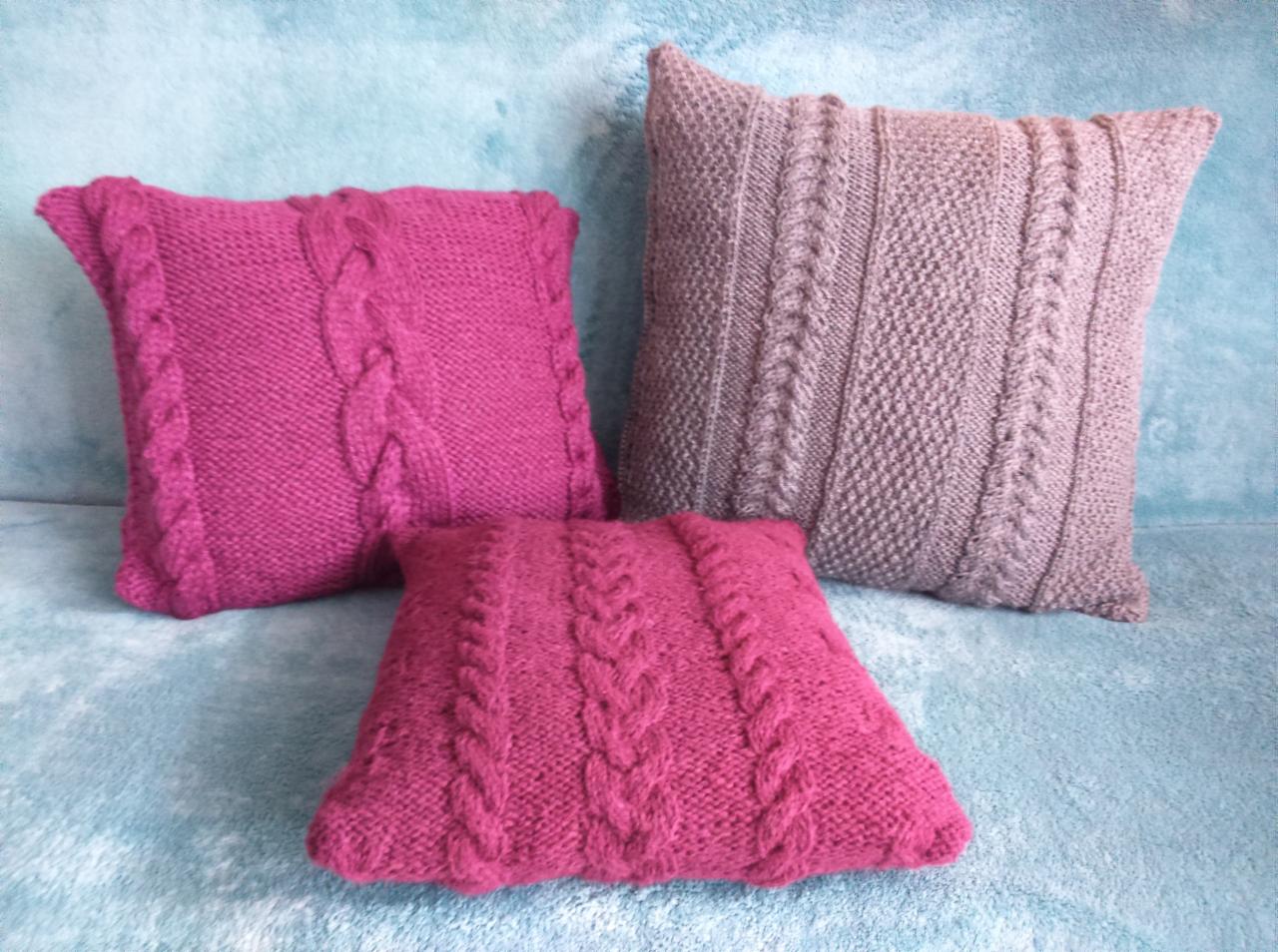 Trzy dziergane poduszki dekoracyjne: dwie różowe z motywem warkocza po bokach i jedna szara z warkoczami pionowymi i wzorem w romby, na tle jasnoniebieskiego pluszowego koca.