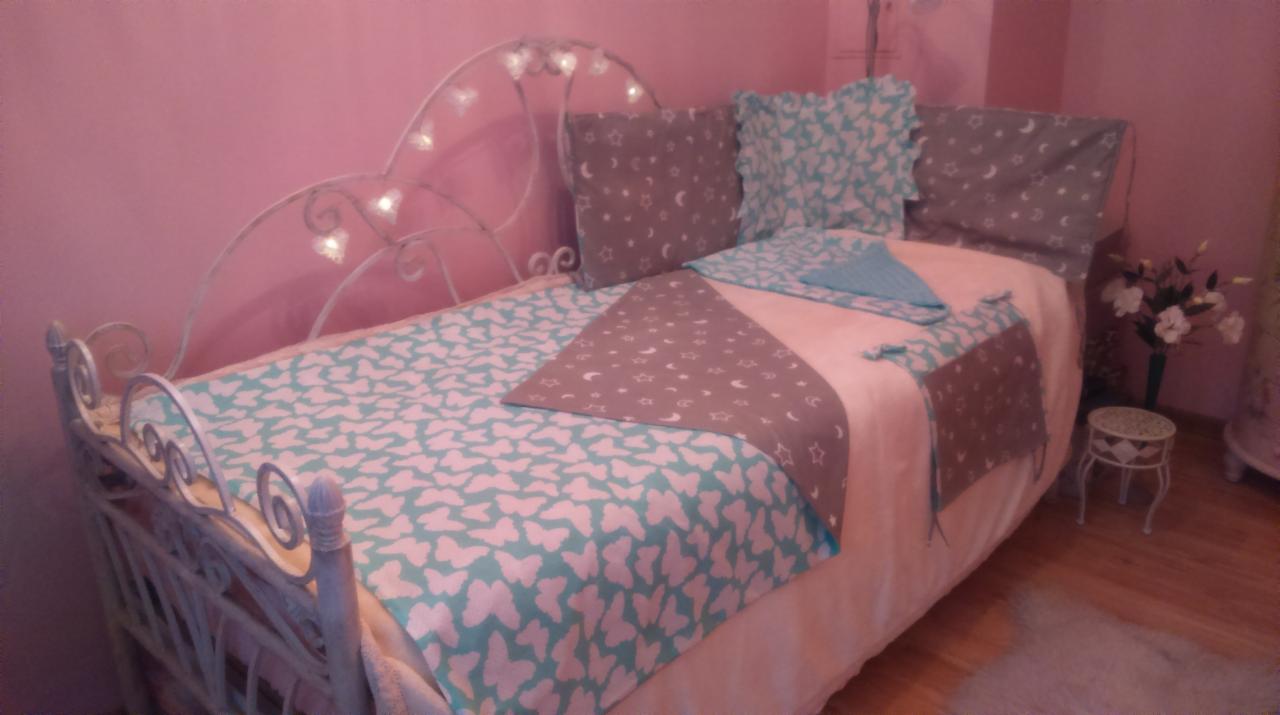 Łóżko dziecięce z metalową ramą, pościel w pastelowych kolorach z motywem motyli i gwiazd, dekoracyjne poduszki, różowe ściany, kwiaty w wazonie.