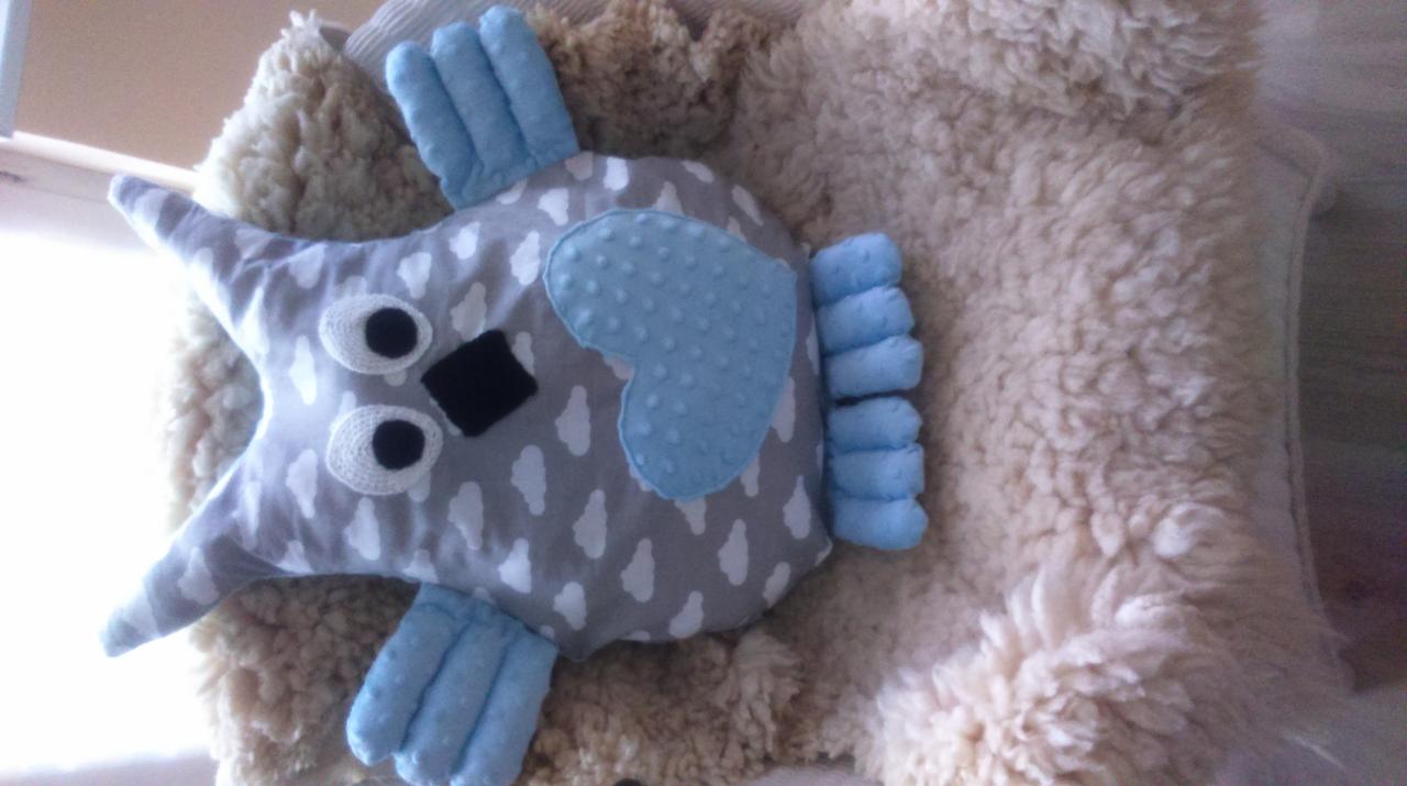 Ręcznie szyta poduszka w kształcie sowy z szarego materiału w białe krople, z niebieskimi elementami: sercem, uszami i brzuchem, leżąca na puszystym, jasnym futrze.