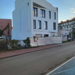 Piotr Jeż, MRÓWEX - Nowoczesny, biały budynek z czarnymi oknami, ogrodzony tymczasowym płotem. Widoczne rusztowania i elementy budowlane. Błękitne niebo w tle.