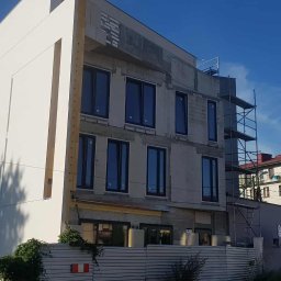 Piotr Jeż, MRÓWEX - Budynek w trakcie ocieplania elewacji, widoczne rusztowania i fragmenty otynkowanej ściany. Część budynku jest już ocieplona i pomalowana na biało. Błękitne niebo w tle.