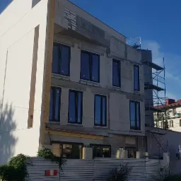 Budynek w trakcie ocieplania elewacji, widoczne rusztowania i fragmenty otynkowanej ściany. Część budynku jest już ocieplona i pomalowana na biało. Błękitne niebo w tle.