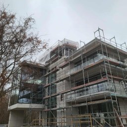 Piotr Jeż, MRÓWEX - Wielopiętrowy budynek w trakcie ocieplania elewacji, otoczony rusztowaniami. Widoczne zabezpieczenia na górnych kondygnacjach. Drzewo z brązowymi liśćmi po lewej stronie.