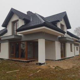 Piotr Jeż, MRÓWEX - Nowoczesny dom jednorodzinny z ciemną dachówką i jasną elewacją w trakcie wykończenia. Widoczne duże okna i elementy konstrukcyjne. Mglista pogoda w tle.