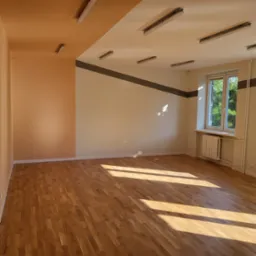 Pomalowane wnętrze pokoju z drewnianą podłogą, grzejnikiem pod oknem i lampami na suficie. Ściany w odcieniach beżu i brzoskwini, z szarym pasem dekoracyjnym.