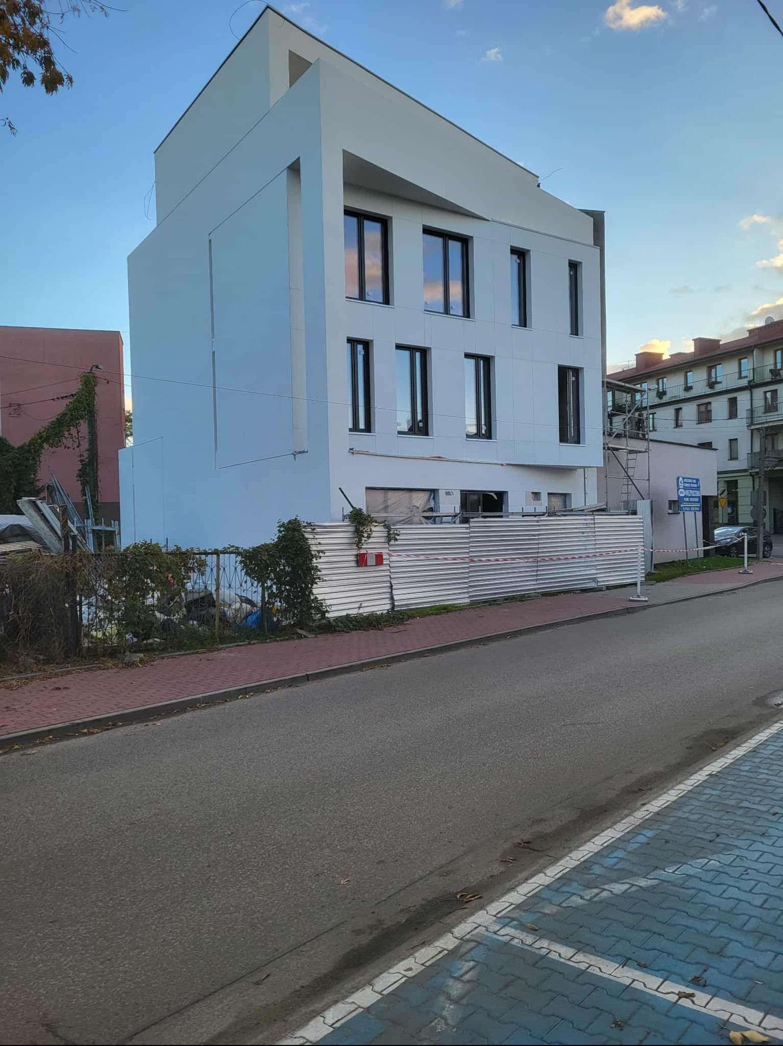 Nowoczesny, biały budynek z czarnymi oknami, ogrodzony tymczasowym płotem. Widoczne rusztowania i elementy budowlane. Błękitne niebo w tle.