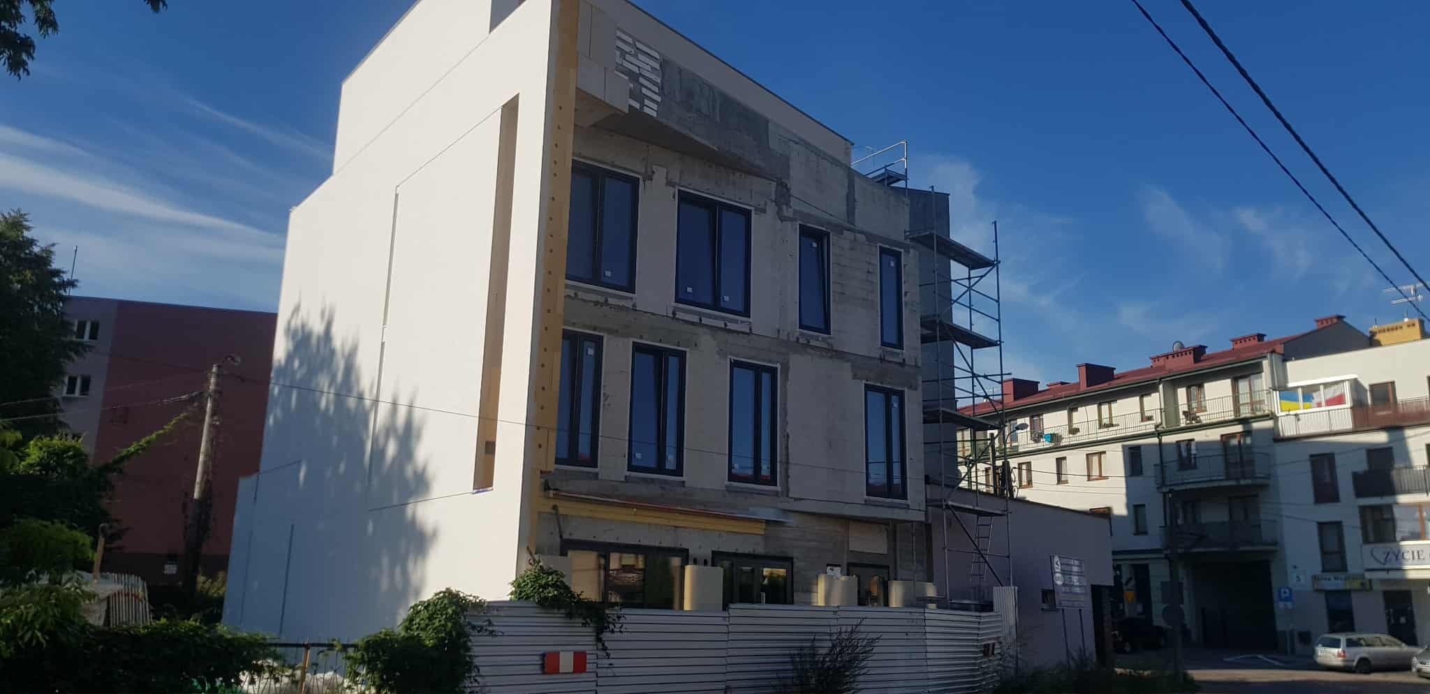 Budynek w trakcie ocieplania elewacji, widoczne rusztowania i fragmenty otynkowanej ściany. Część budynku jest już ocieplona i pomalowana na biało. Błękitne niebo w tle.