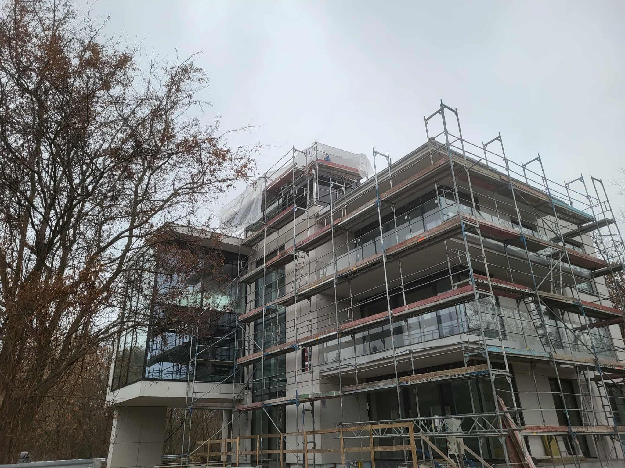 Wielopiętrowy budynek w trakcie ocieplania elewacji, otoczony rusztowaniami. Widoczne zabezpieczenia na górnych kondygnacjach. Drzewo z brązowymi liśćmi po lewej stronie.