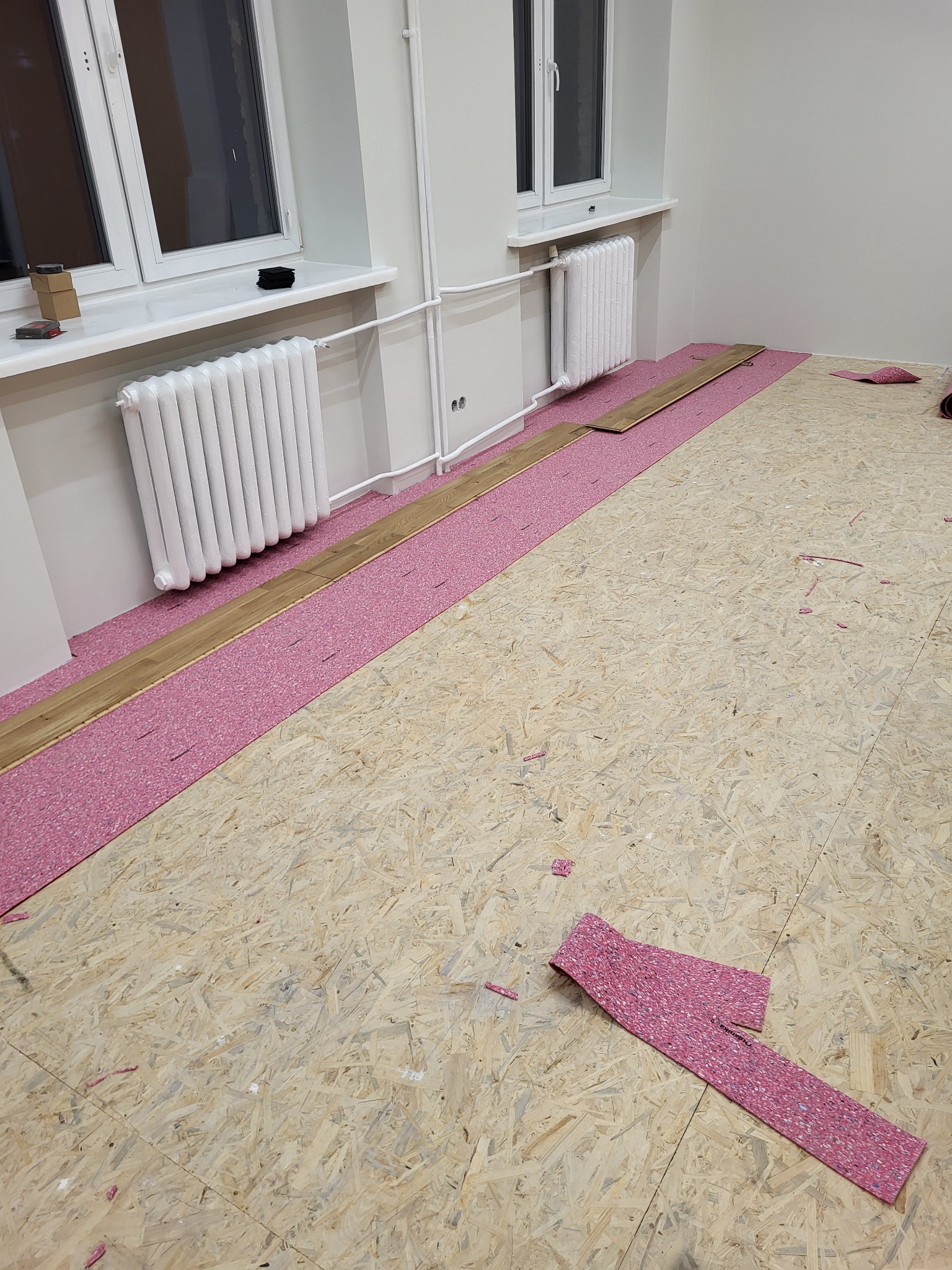 Montaż podłogi: płyta OSB, różowa pianka i panele. Widoczne grzejniki i okna. Etap prac przed ułożeniem całej podłogi. Jasne wnętrze, neutralne kolory ścian.