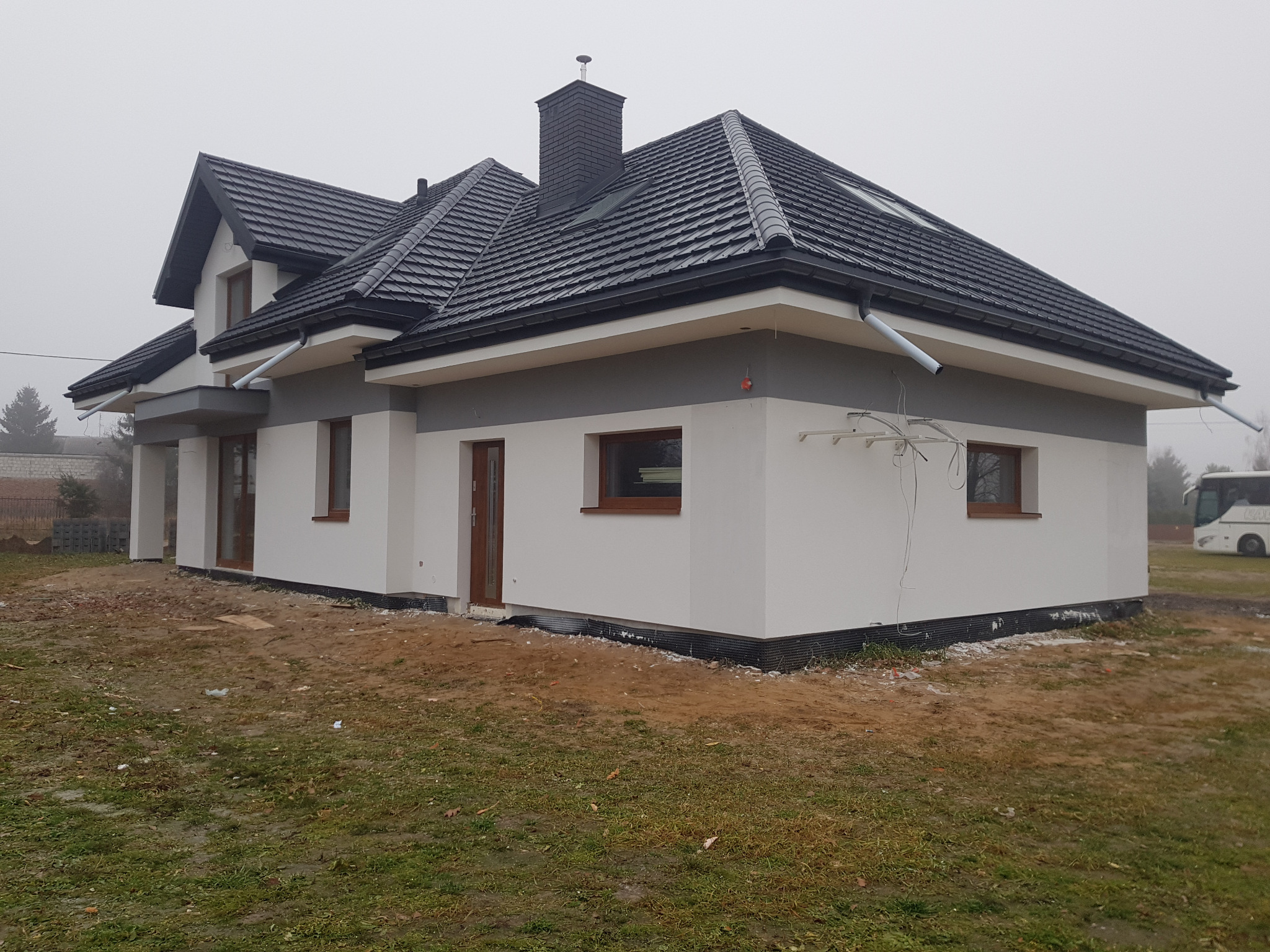 Nowoczesny dom jednorodzinny z ciemną dachówką i jasną elewacją w pochmurny dzień. Widoczne detale wykończenia i otoczenie budynku.