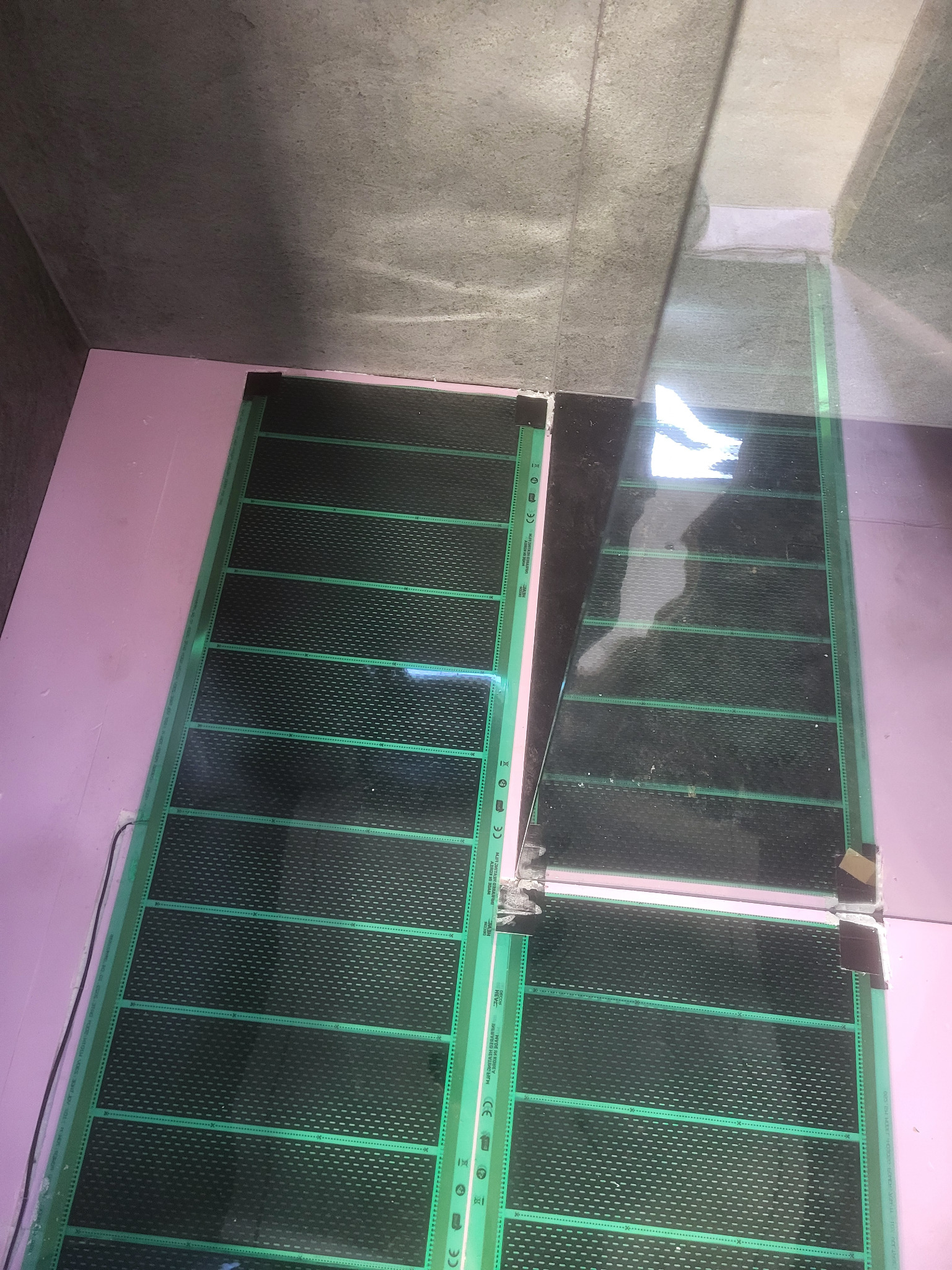 Rozłożone maty grzewcze na różowej izolacji podłogowej, przygotowane do zalania wylewką. Widoczne kable i łączenia mat. Surowy beton na ścianach i suficie.