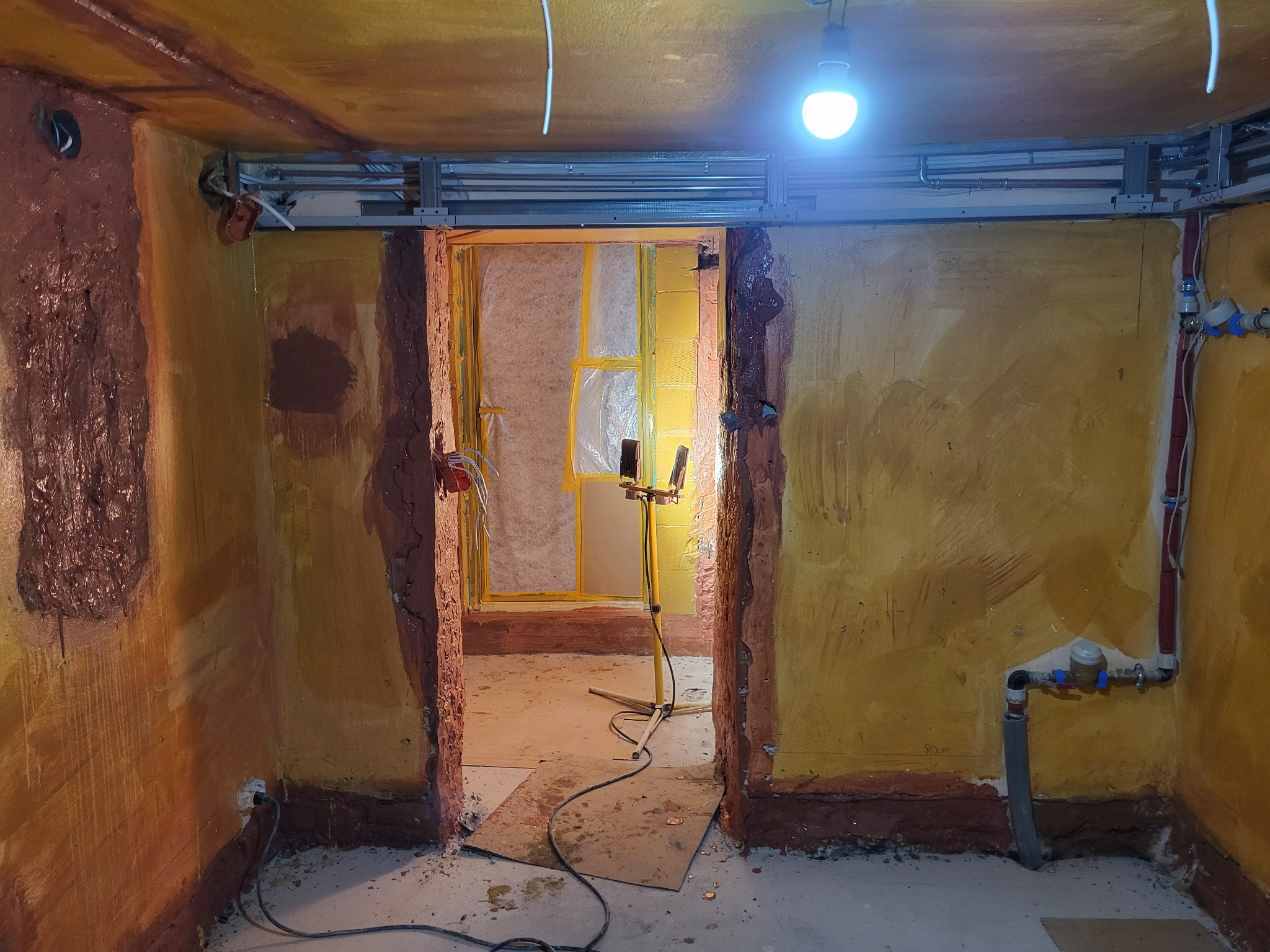 Pokój w trakcie prac wykończeniowych: surowe ściany w odcieniach żółci i brązu, widoczne instalacje, zabezpieczone folią drzwi, oświetlenie robocze.