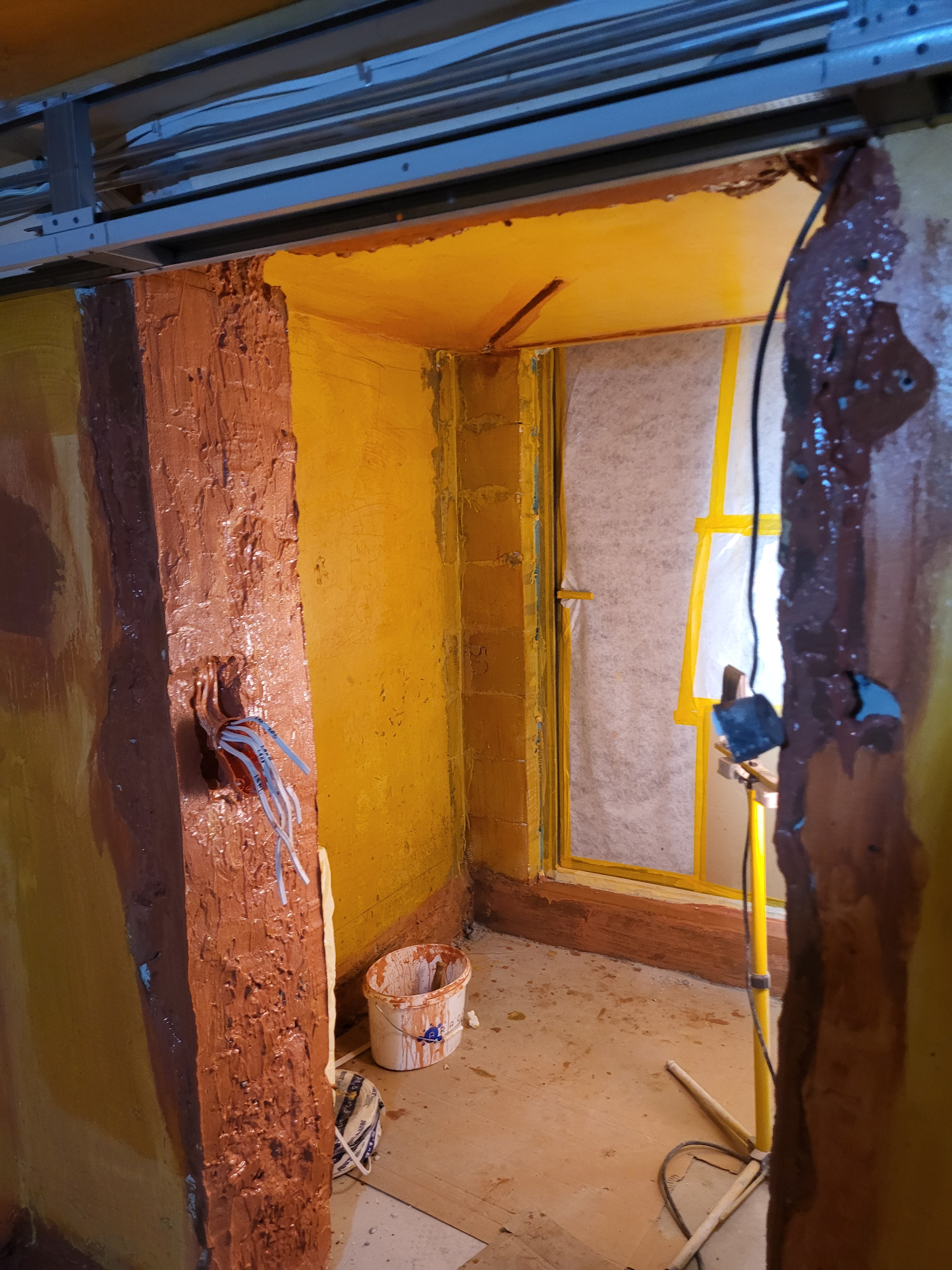 Żółto-brązowe wnętrze w trakcie prac wykończeniowych. Widoczne kable, wiadro z farbą i lampa. Ściany częściowo pokryte brązową substancją, okno zabezpieczone folią.