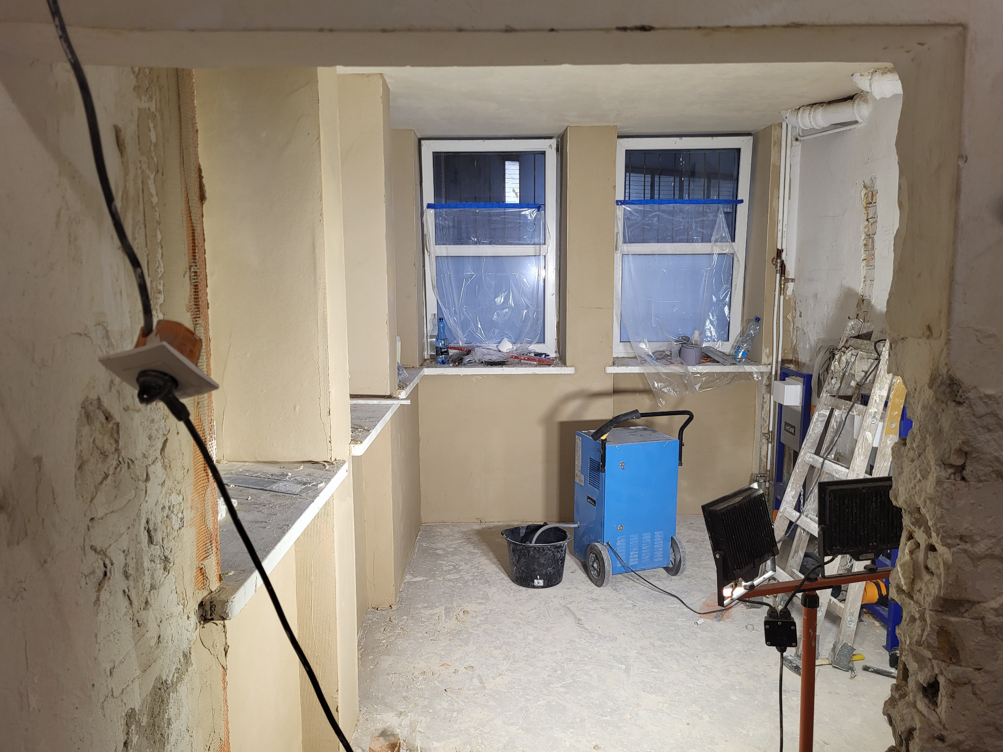 Pokój w trakcie prac wykończeniowych: ściany z nałożoną gładzią, zabezpieczone okna folią, sprzęt budowlany, widoczne ślady po kuciu. Fragment ściany z odsłoniętą cegłą.