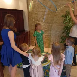 Dzieci obserwują tworzenie gigantycznej bańki mydlanej przez osobę dorosłą w pomieszczeniu z dekoracyjną ścianą z imitacji kamienia.