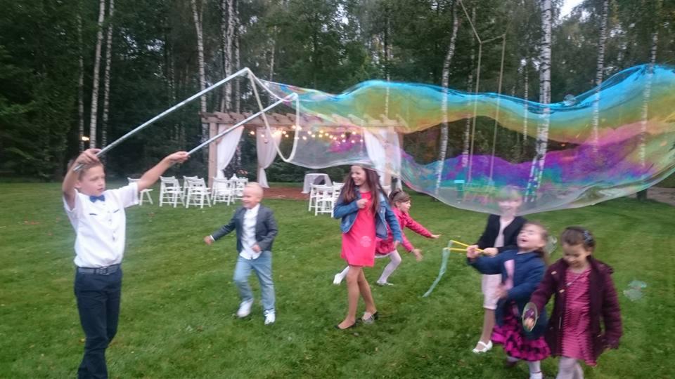 Chłopiec w białej koszuli robi ogromne bańki mydlane za pomocą specjalnego sznurka, wokół niego na zielonym trawniku bawią się dzieci, w tle drewniana altana z białymi zasłonami.