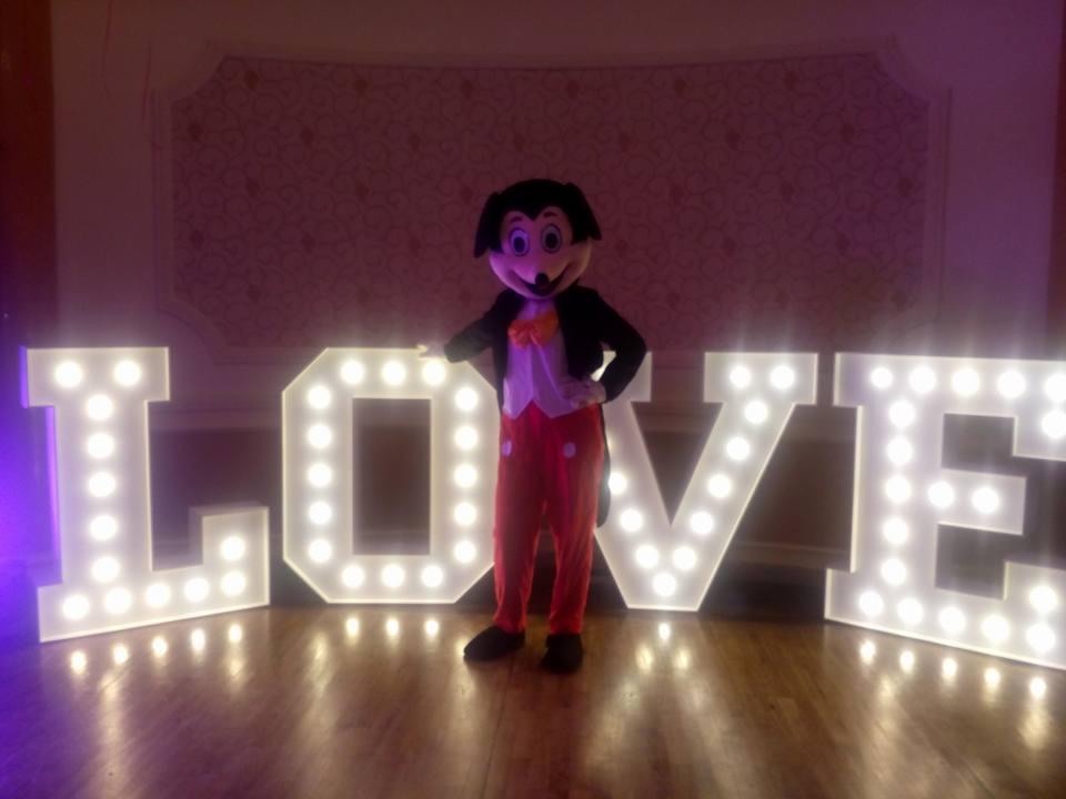 Osoba w kostiumie postaci z kreskówki stoi za podświetlanym napisem LOVE na drewnianej podłodze, z wzorzystą ścianą w tle.