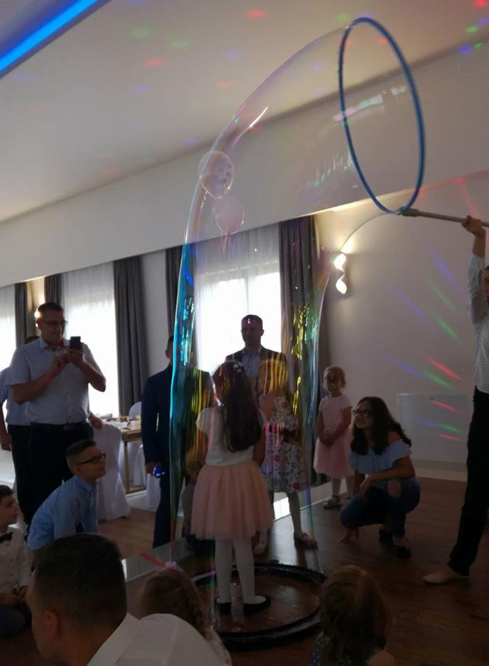 Dzieci obserwują tworzenie ogromnych baniek mydlanych podczas imprezy, oświetlenie dyskotekowe na suficie.
