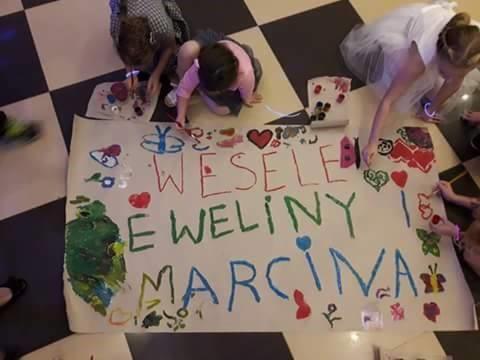 Dzieci malują farbami na dużym arkuszu papieru życzenia dla Eweliny i Marcina z okazji wesela, na czarno-białej podłodze.