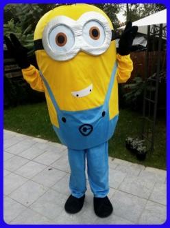 Żółty kostium Minionka z podniesionymi rękami i szerokim uśmiechem, stojący na jasnych płytkach chodnikowych z zielonym tłem.