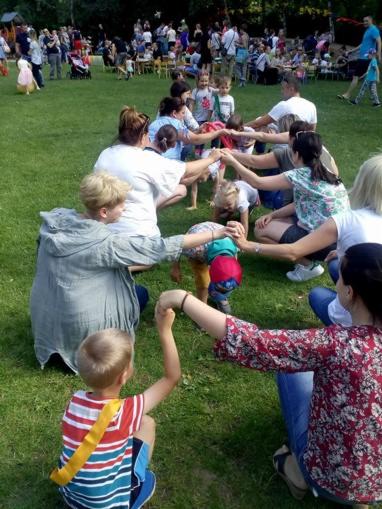 Grupa dzieci i dorosłych trzymających się za ręce, tworzących tunel na trawie podczas zabawy na świeżym powietrzu, w tle widoczny tłum ludzi.