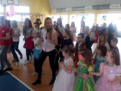 Kobieta z mikrofonem prowadzi zabawę dla grupy dzieci w sali gimnastycznej, widoczne dekoracje balonowe i efekty świetlne.