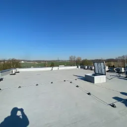 Płaski dach budynku z zainstalowanymi klimatyzatorami, systemem odgromowym i otwartym włazem. W tle widok na zielone tereny i błękitne niebo.