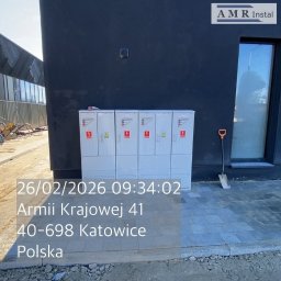 AMR Instal - Sześć białych szafek rozdzielczych z czerwonymi symbolami ostrzegawczymi, zamontowanych na ciemnej elewacji budynku obok okna, z łopatą opartą o ścianę.