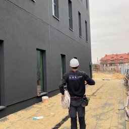 AMR Instal - Pracownik w białym kasku i stroju 'AMR Instal' niesie zwinięte białe przewody, idąc wzdłuż nowoczesnego budynku z ciemnoszarą elewacją. Na ziemi piasek i rynna odwadniająca.