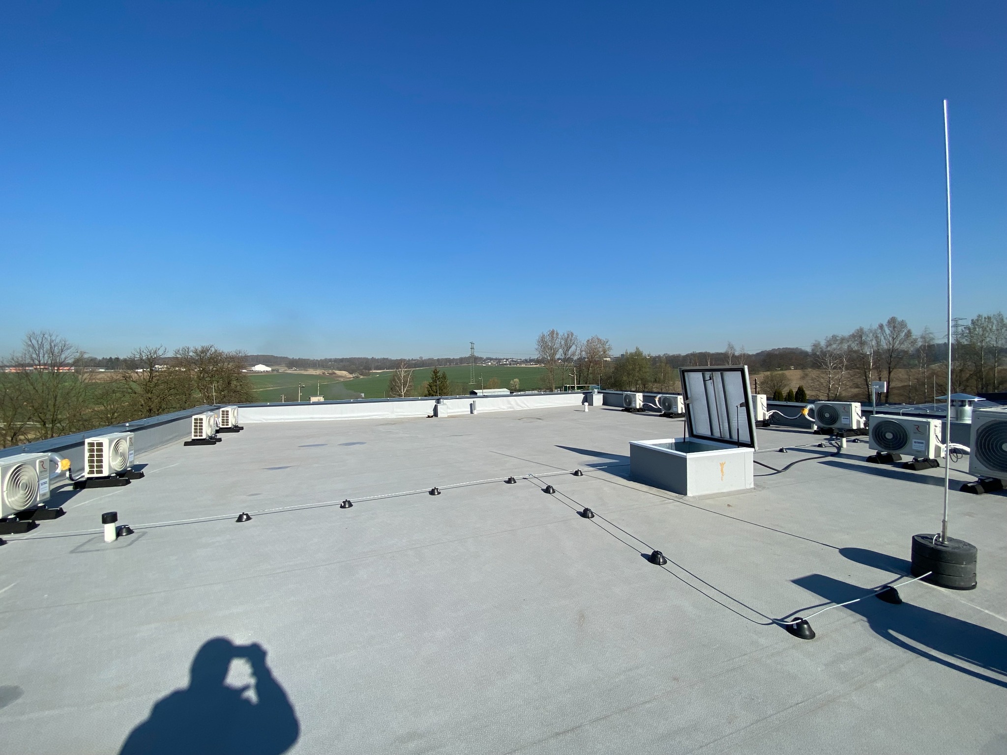 Płaski dach budynku z zainstalowanymi klimatyzatorami, systemem odgromowym i otwartym włazem. W tle widok na zielone tereny i błękitne niebo.