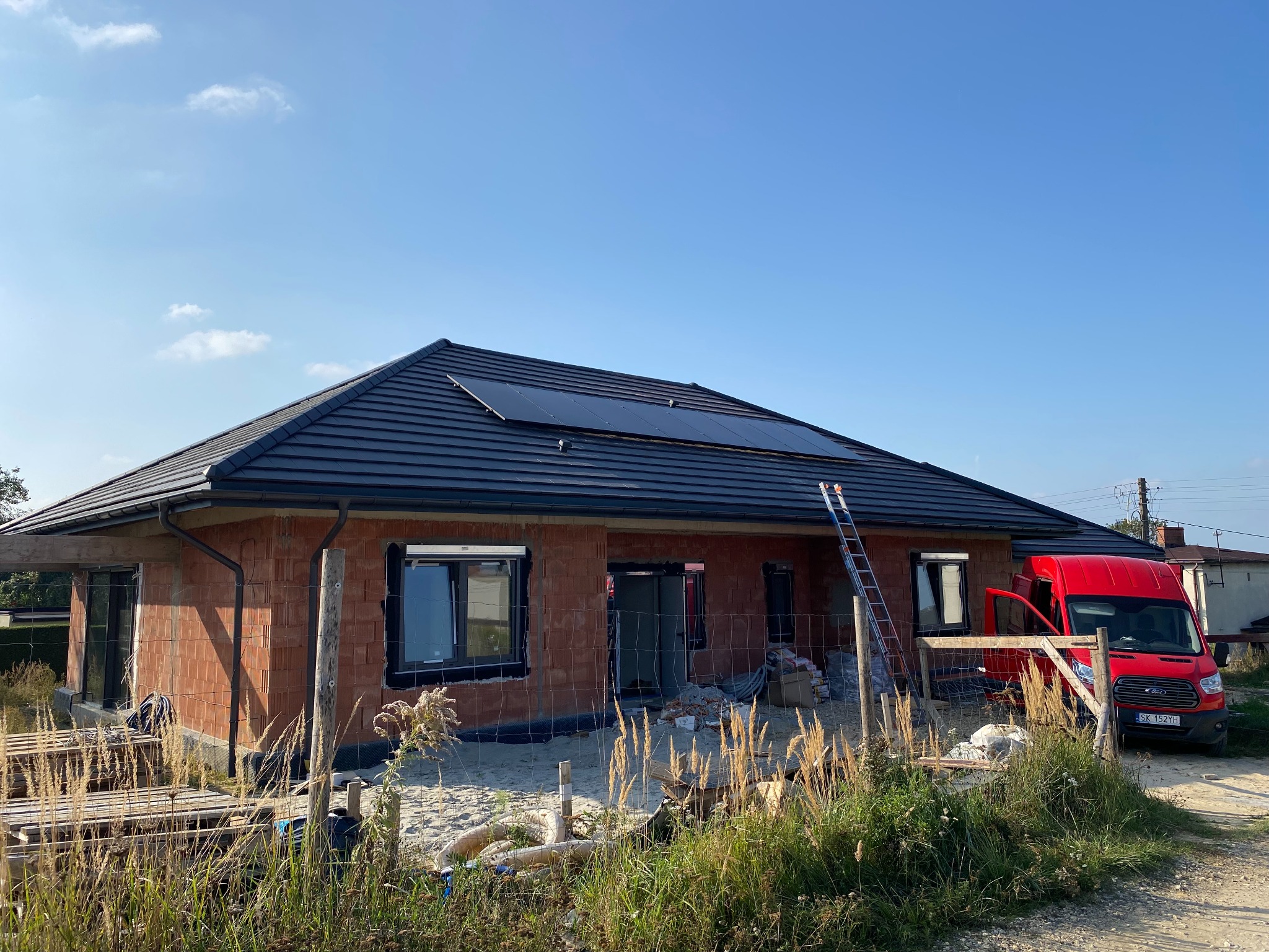 Dom w trakcie budowy z panelami fotowoltaicznymi na dachu, widoczna drabina i czerwony samochód dostawczy. Stan surowy otwarty, cegła.