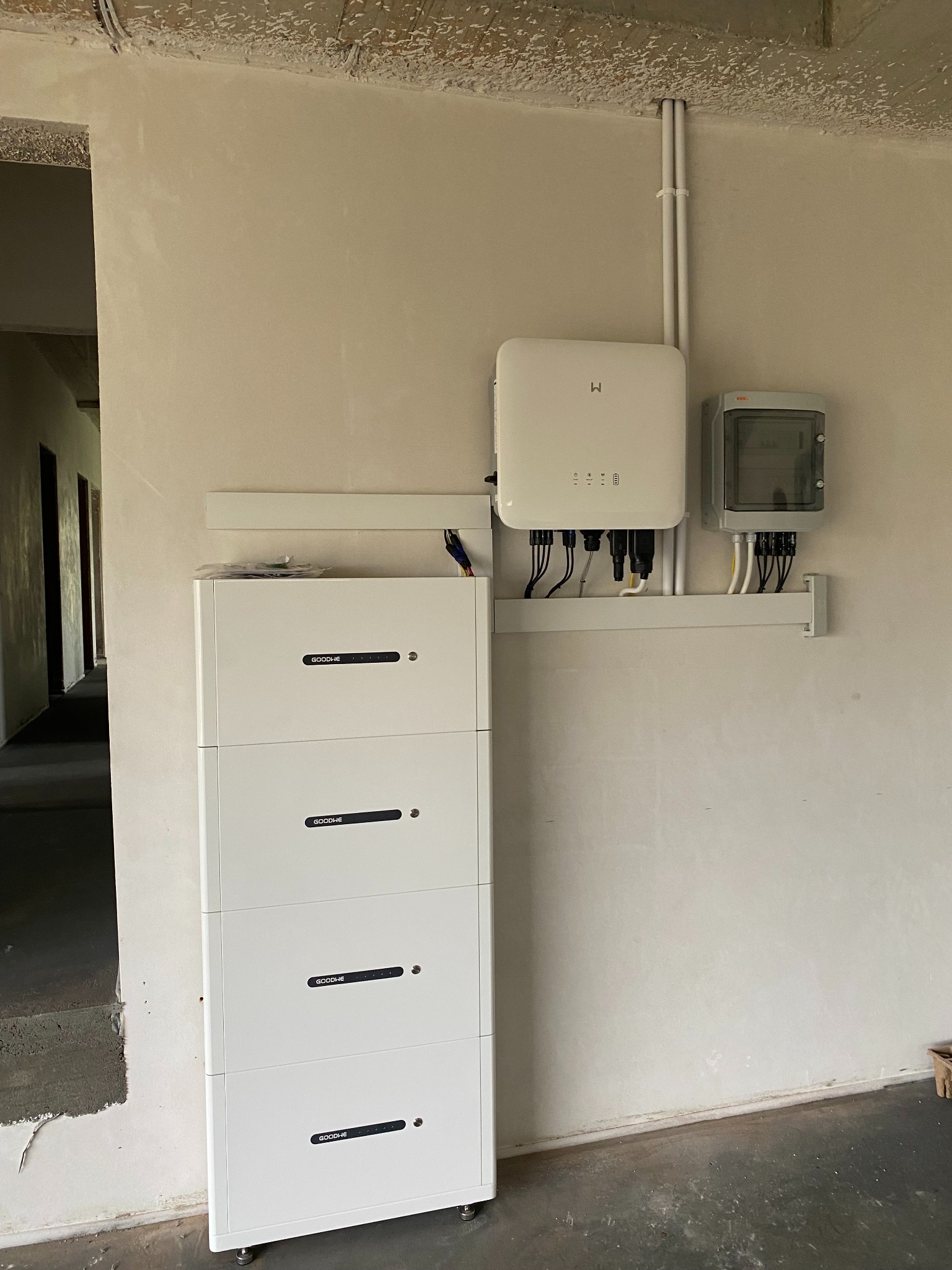 Biały akumulator energii GoodWe, inwerter i skrzynka elektryczna zamontowane na ścianie w surowym wnętrzu. Widoczne kable i przewody. Instalacja fotowoltaiczna w trakcie montażu.