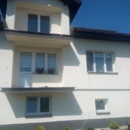 Jasna, dwukondygnacyjna elewacja domu jednorodzinnego z oknami z roletami dzień-noc i balkonem, z widocznymi elementami wykończeniowymi i fragmentem dachu, sąsiedni budynek w tle.