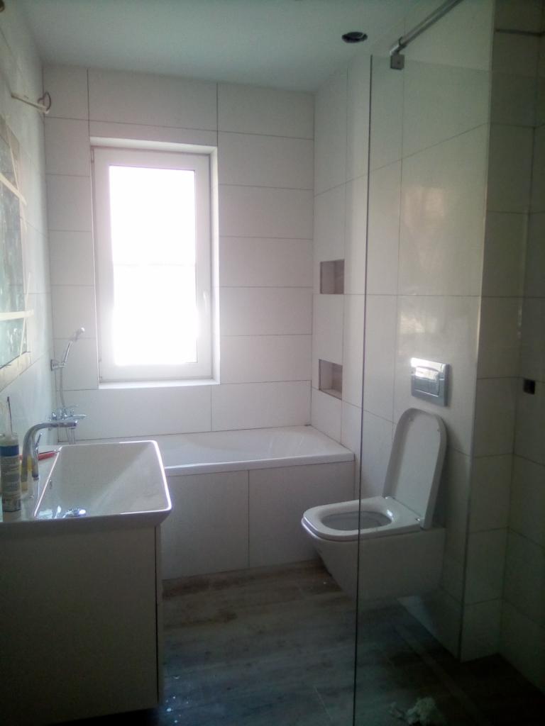 Wykończona łazienka z białymi płytkami, umywalką z szafką, zabudowaną wanną i podwieszaną toaletą, widoczne szklane drzwi kabiny prysznicowej.