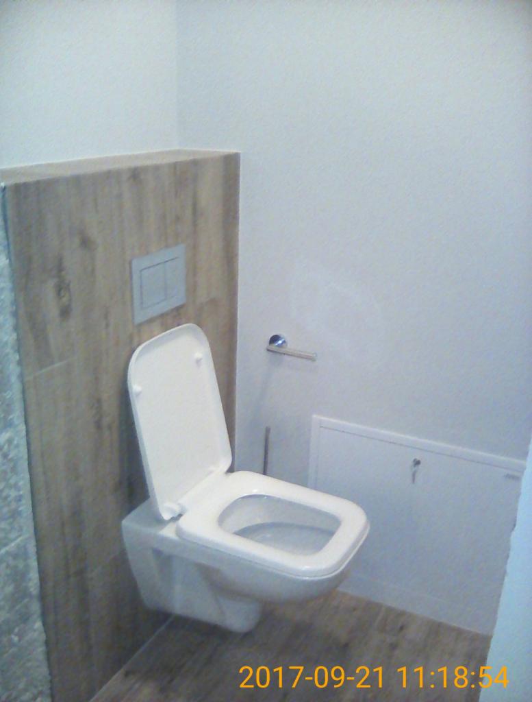 Wisząca toaleta z deską podniesioną do góry, zamontowana przy obudowie ze wzorem drewna, z widocznym przyciskiem spłukiwania i drzwiczkami rewizyjnymi w tle.