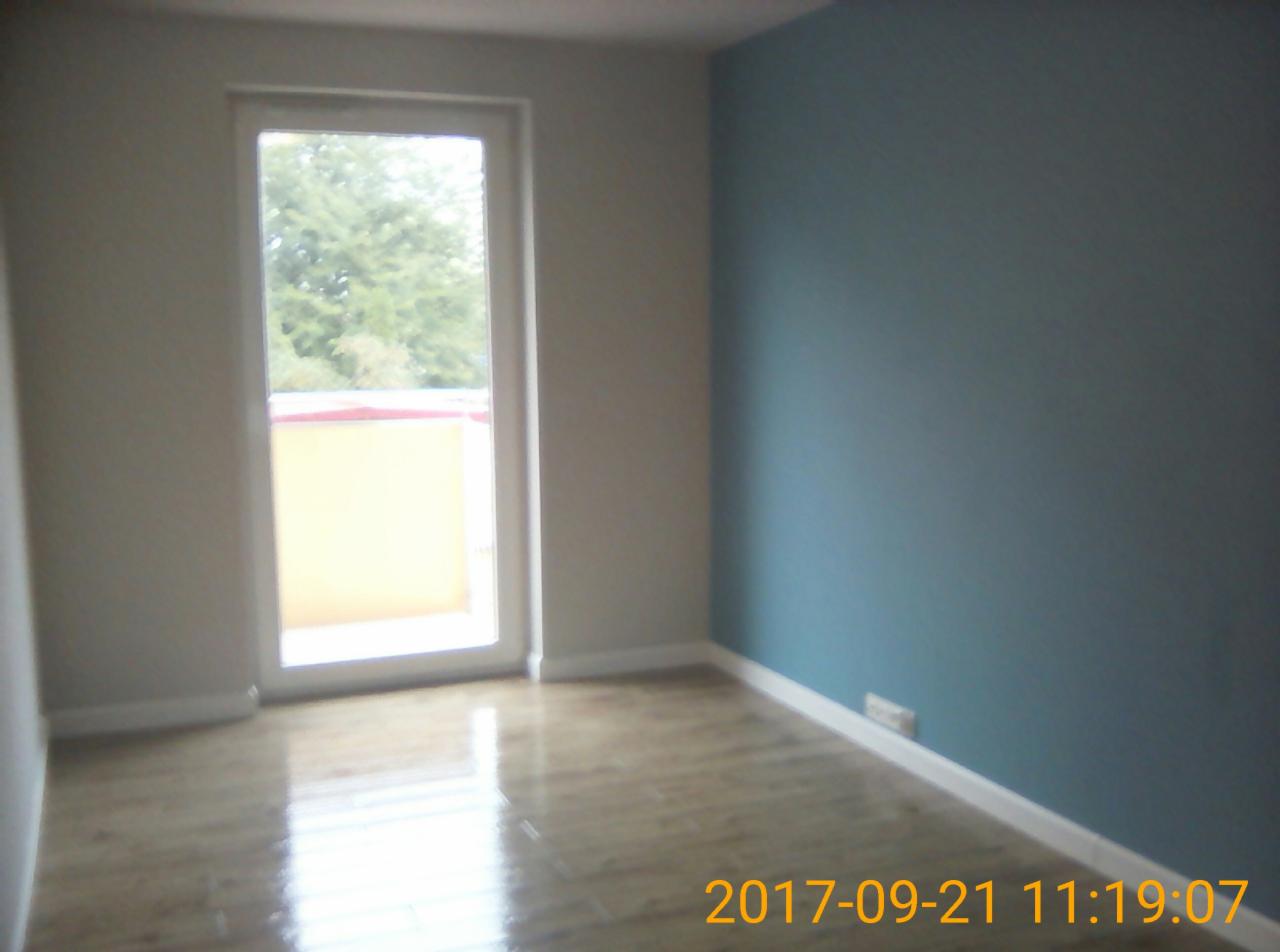 Pomalowane na niebiesko ściany w pustym pokoju z parkietem i białymi listwami przypodłogowymi, okno z widokiem na zieleń.