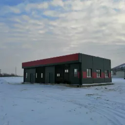 Nowoczesny budynek z płyt warstwowych w zimowym krajobrazie, widoczne ślady opon na śniegu, w tle dom jednorodzinny.