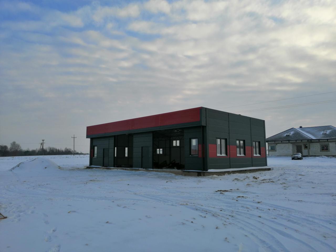 Nowoczesny budynek z płyt warstwowych w zimowym krajobrazie, widoczne ślady opon na śniegu, w tle dom jednorodzinny.