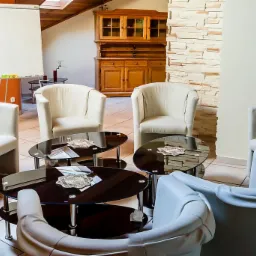 Jasne fotele ustawione w kręgu wokół niskich, okrągłych stolików z ciemnymi blatami, na których leżą ozdobne serwetki. W tle widoczna tablica suchościeralna, drewniana komoda i fragment ściany...