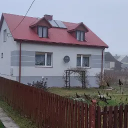 Dom jednorodzinny po remoncie elewacji i ociepleniu, z czerwoną blachodachówką i panelami słonecznymi na dachu, widoczny zza drewnianego płotu w mglisty dzień.