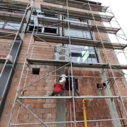 Salon Buderus TOP PARTNER - Murarz w czerwonej kurtce montuje elementy wentylacji na rusztowaniu przy ceglanym budynku z oknami.