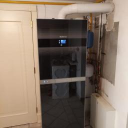 Salon Buderus TOP PARTNER - Nowoczesny, czarny panel grzewczy Buderus zamontowany w pomieszczeniu z widocznymi rurami i instalacją hydrauliczną, obok białe drzwi.