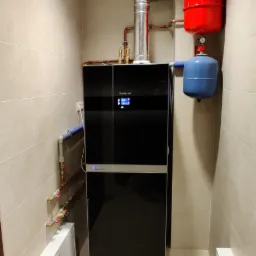 Kompaktowa kotłownia: czarny piec Buderus z podłączeniami miedzianymi i zbiornikami wyrównawczymi w kolorach czerwonym i niebieskim, umieszczona w pomieszczeniu wyłożonym jasnymi płytkami.