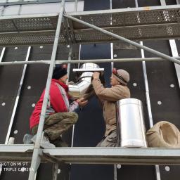 Salon Buderus TOP PARTNER - Montaż stalowego komina systemowego przez dwóch monterów na rusztowaniu w Raszynie, widoczne ocieplenie budynku i elementy mocujące.