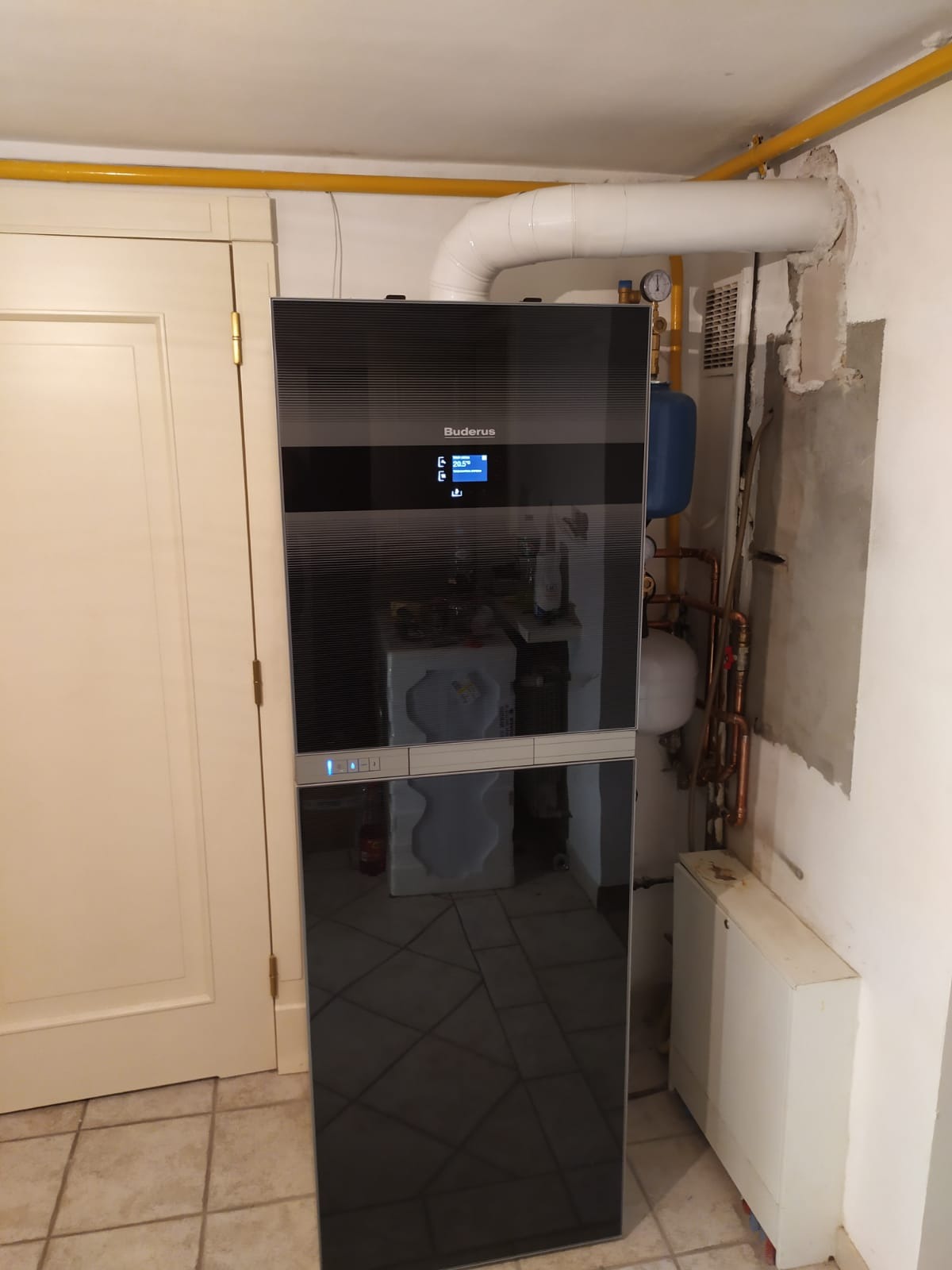 Nowoczesny, czarny panel grzewczy Buderus zamontowany w pomieszczeniu z widocznymi rurami i instalacją hydrauliczną, obok białe drzwi.