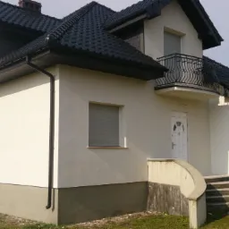 Dwupiętrowy dom z białymi ścianami, czarnym dachem i balkonem z ozdobną, czarną balustradą, widok z boku, częściowo ocieniony, z fragmentem osoby w tle.