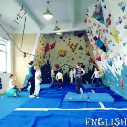 Dzieci w sali zabaw z kolorową ścianką wspinaczkową, niebieską podłogą i materacami asekuracyjnymi, jedno dziecko wspina się na ściankę, inne stoją na podłodze, a dziewczynka siedzi na zielonej...