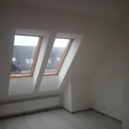 Poddasze z dwoma oknami dachowymi, białe ściany po świeżym wykończeniu, szary panel podłogowy, grzejnik pod oknami.