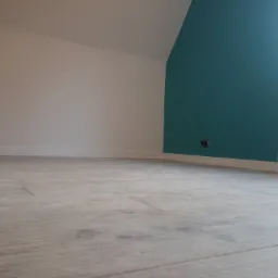 Pokój po remoncie: świeżo ułożone panele podłogowe w odcieniach szarości, białe ściany przechodzące w skośny sufit, fragment ściany pomalowany na turkusowo, widoczny grzejnik i gniazdko elektryczne.